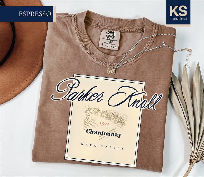 a brown t-shirt with a label that reads "Parker Knoll 1994 Chardonnay Napa Valley" and a small tag that says "Espresso". The t-shirt is laid out on a white surface, with a brown hat and some leaves visible in the background.