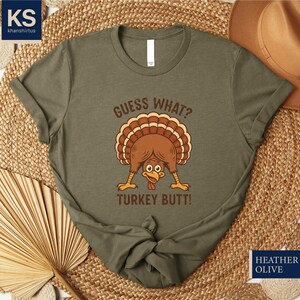 A green t-shirt with a humorous graphic of a turkey and the text "Guess What? Turkey Butt!" printed on it, along with a straw hat and a woven mat in the background.