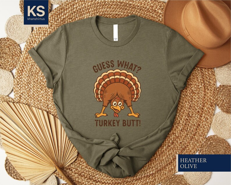 A green t-shirt with a humorous graphic of a turkey and the text "Guess What? Turkey Butt!" printed on it, along with a straw hat and a woven mat in the background.