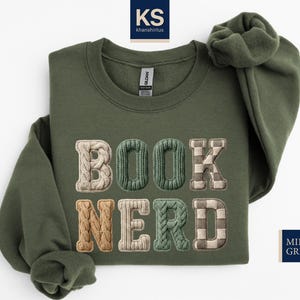 May include: Olive green sweatshirt with the words "BOOK NERD" embroidered in textured, multi-colored letters. The letters have a knitted or plaid appearance. The sweatshirt is folded, with the sleeves visible. The text "MILITARY GREEN" is in a navy blue rectangle.