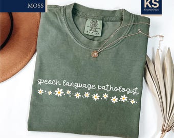 May include: Moss green t-shirt with the words "Speech language pathologist" in white script, decorated with white daisy graphics. The shirt is folded, with a gold necklace and a brown hat visible.
