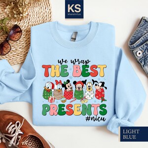 A light blue t-shirt with a colorful graphic design featuring cartoon characters and the text "we wrap the best presents #nuc" is displayed on a white background.
