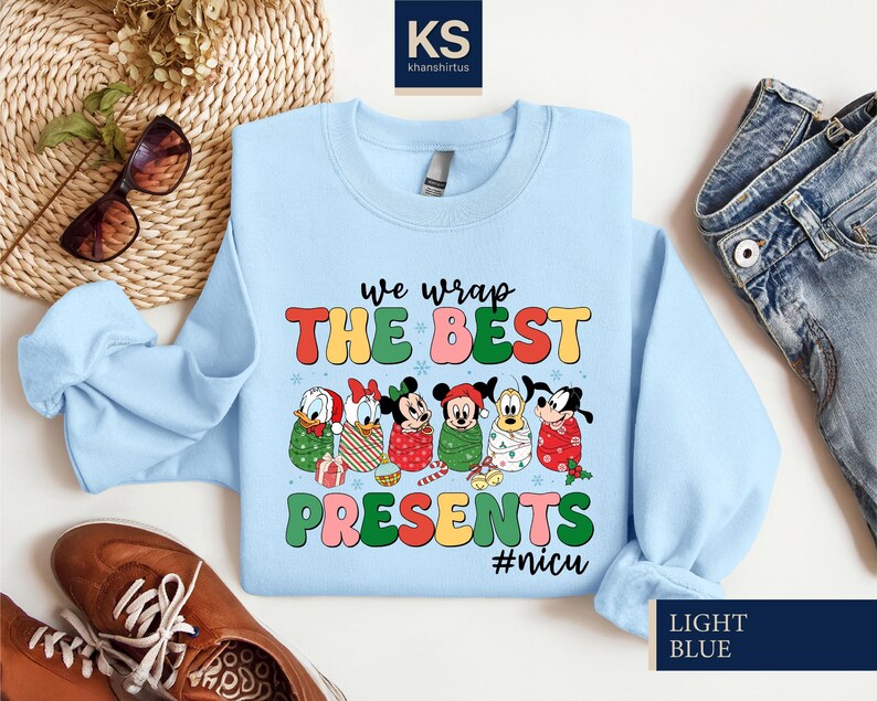 A light blue t-shirt with a colorful graphic design featuring cartoon characters and the text "we wrap the best presents #nuc" is displayed on a white background.