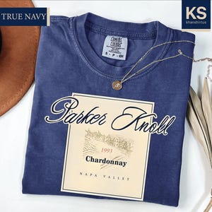a navy blue t-shirt with a wine label design, a brown hat, and some dried leaves, all placed on a white background.