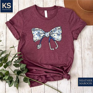 May include: A maroon t-shirt with a white and blue floral bow and a stethoscope design. The text "HEATHER MAROON" is printed on the bottom right corner of the image.