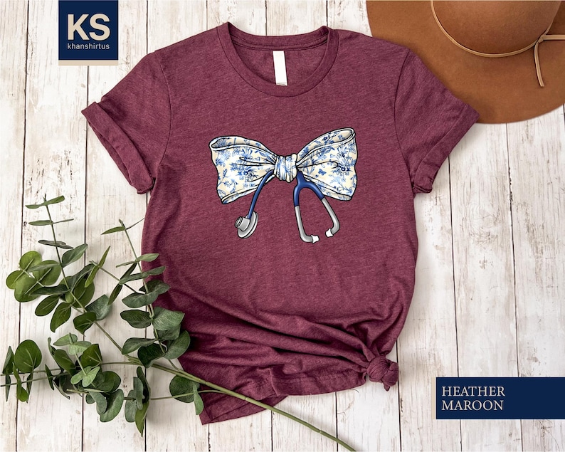 May include: A maroon t-shirt with a white and blue floral bow and a stethoscope design. The text "HEATHER MAROON" is printed on the bottom right corner of the image.