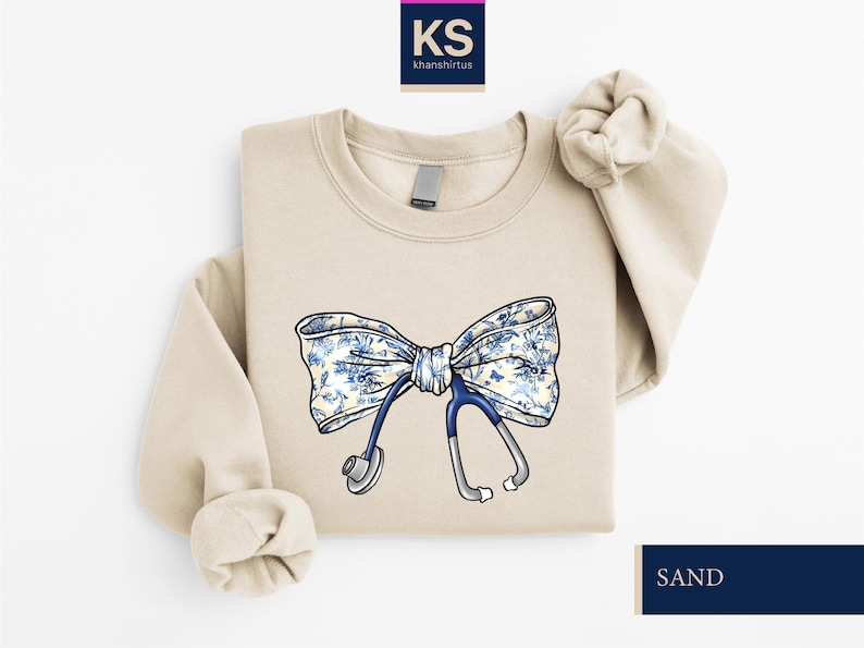 May include: A beige sweatshirt with a blue and white floral bow and a stethoscope. The text 'KS khanshirtus' is in the top left corner of the image. The text 'SAND' is in the bottom right corner of the image.