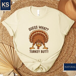 A beige t-shirt with a humorous graphic of a turkey and the text "Guess what? Turkey butt!" printed on it, along with a straw hat and a woven mat in the background.