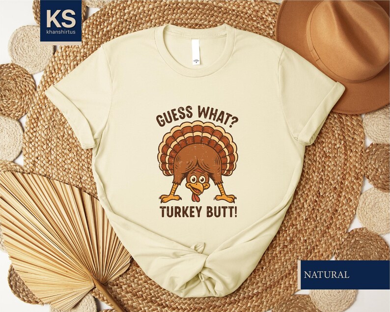 A beige t-shirt with a humorous graphic of a turkey and the text "Guess what? Turkey butt!" printed on it, along with a straw hat and a woven mat in the background.