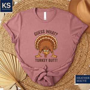 A pink t-shirt with a humorous graphic of a turkey and the text "Guess What? Turkey Butt!" printed on it, along with a straw hat and a woven mat in the background.