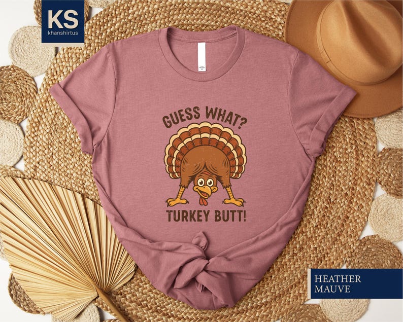 A pink t-shirt with a humorous graphic of a turkey and the text "Guess What? Turkey Butt!" printed on it, along with a straw hat and a woven mat in the background.