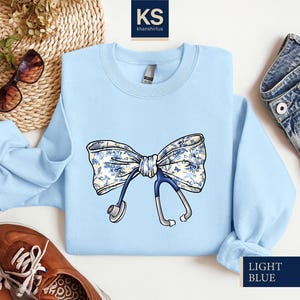 May include: Light blue sweatshirt with a white and blue floral bow and a stethoscope. The text "LIGHT BLUE" is printed on the bottom right corner of the sweatshirt.