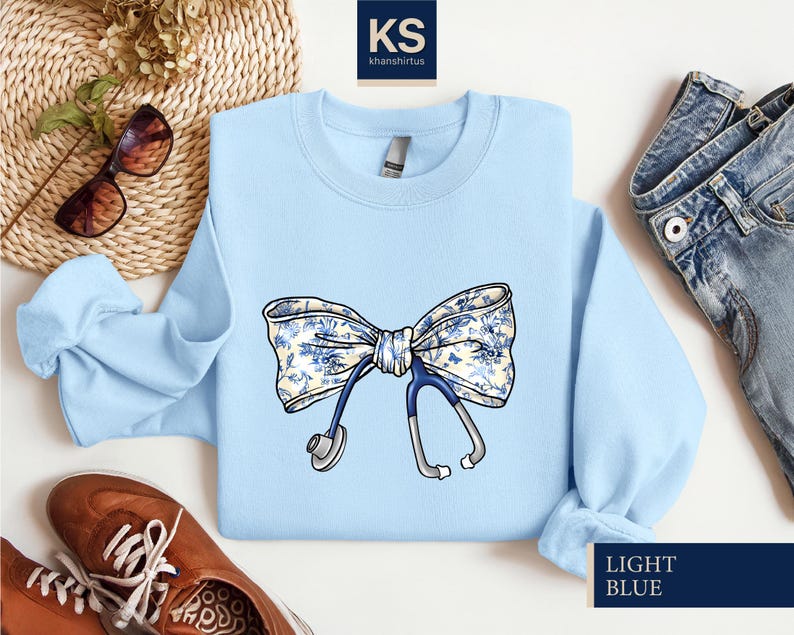 May include: Light blue sweatshirt with a white and blue floral bow and a stethoscope. The text "LIGHT BLUE" is printed on the bottom right corner of the sweatshirt.