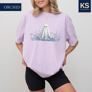 A woman wearing a pink t-shirt with a graphic of a ghost standing in a field of flowers.