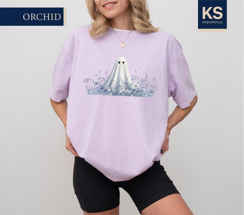 A woman wearing a pink t-shirt with a graphic of a ghost standing in a field of flowers.
