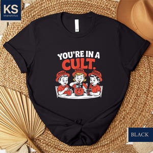 May include: Black t-shirt with a graphic of three children wearing red hats with the word "MAGA" on them. The text "YOU'RE IN A CULT." is above the children.