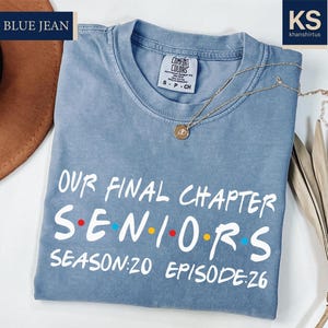 May include: A light blue t-shirt with the text "OUR FINAL CHAPTER SENIORS SEASON:20 EPISODE:26" in white. The shirt has a crew neck and short sleeves. A gold necklace with a pendant is on the shirt.
