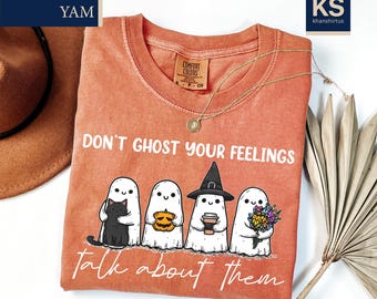 May include: An orange Comfort Colors t-shirt with the slogan "DON'T GHOST YOUR FEELINGS talk about them." The shirt displays a graphic of four cartoon ghosts, each holding a different item: a black cat, a pumpkin, a coffee cup, and flowers.