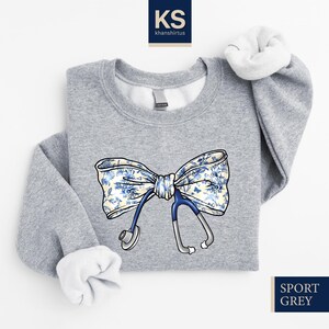 May include: A gray sweatshirt with a blue stethoscope and a white bow with a blue floral pattern. The text "SPORT GREY" is in the bottom right corner of the image.