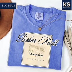 A blue t-shirt with a wine label printed on it, along with a brown hat and some dried leaves, on a white background.