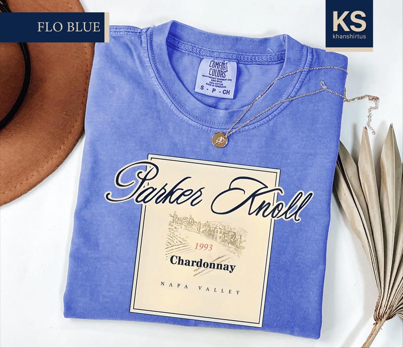 A blue t-shirt with a wine label printed on it, along with a brown hat and some dried leaves, on a white background.