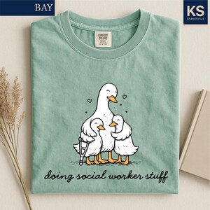 May include: Sage green t-shirt with a graphic of a mother duck and two ducklings hugging. The mother duck has a crutch. The text "doing social worker stuff" is printed below the graphic. The shirt has a crew neck and short sleeves.
