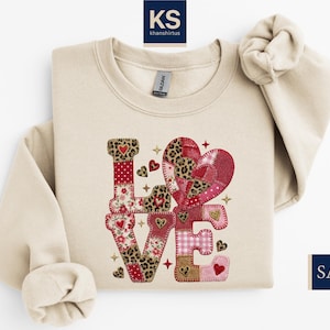 May include: A sand-colored sweatshirt with the word "LOVE" appliqued on the front. The letters are made of various patterned fabrics, including leopard print, floral, and hearts. The design is embellished with small hearts and stars.