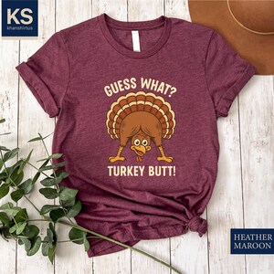 A burgundy t-shirt with a humorous graphic of a turkey and the text "Guess What? Turkey Butt!" is displayed on a wooden surface, accompanied by a brown hat and some greenery.