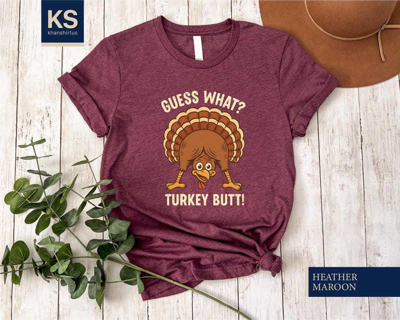 A burgundy t-shirt with a humorous graphic of a turkey and the text "Guess What? Turkey Butt!" is displayed on a wooden surface, accompanied by a brown hat and some greenery.