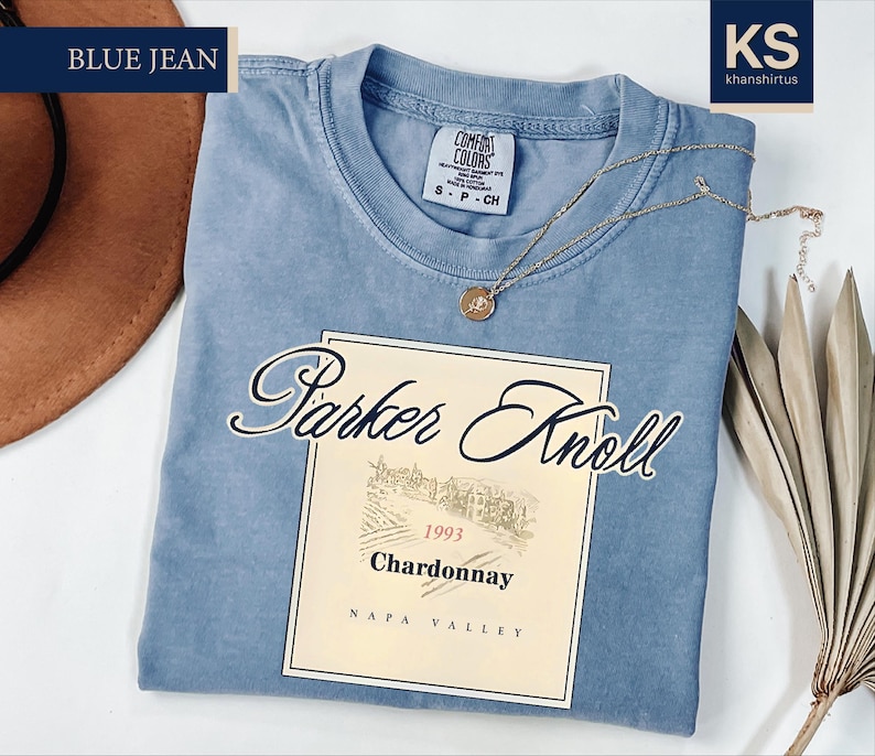 a blue t-shirt with a wine label design, a hat, and some dried leaves, all placed on a white background.