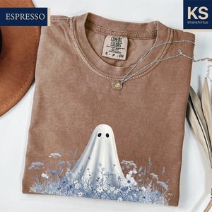 a brown t-shirt with a ghost design printed on it, along with a hat and some dried leaves.