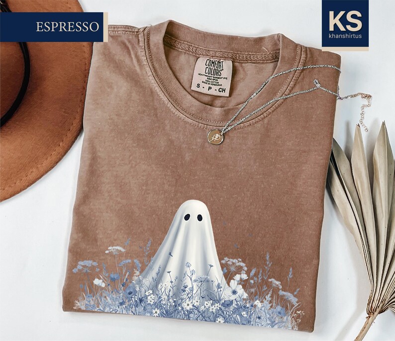 a brown t-shirt with a ghost design printed on it, along with a hat and some dried leaves.