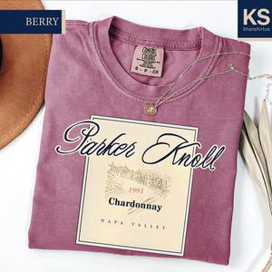 a pink t-shirt with a wine label design, a hat, and some dried leaves, all placed on a white background.