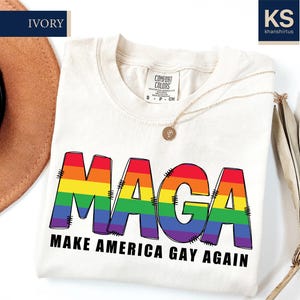 May include: Ivory-colored t-shirt with the word "MAGA" in rainbow colors and the text "MAKE AMERICA GAY AGAIN" in black. The shirt has a crew neck and short sleeves. A gold necklace is draped across the shirt.