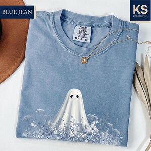 a blue t-shirt with a ghost design, a brown hat, and some dried leaves, all placed on a white surface.