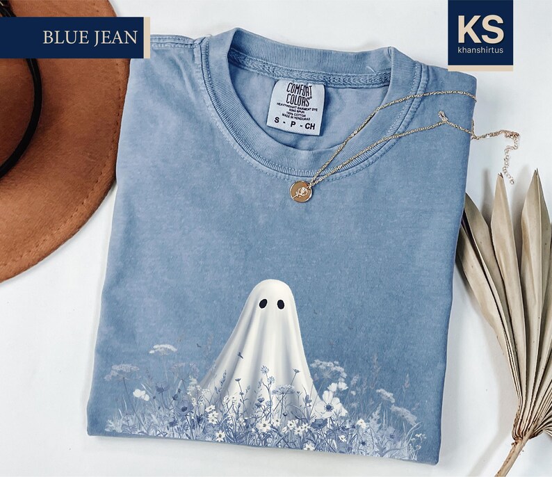 a blue t-shirt with a ghost design, a brown hat, and some dried leaves, all placed on a white surface.