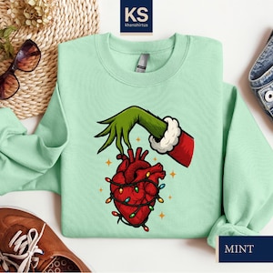 A mint green sweatshirt with a graphic of a hand holding a heart with lights, a Santa Claus hand, and a Christmas tree. The sweatshirt is laid out on a white surface, along with a pair of sunglasses, a pair of jeans, and a pair of brown shoes.