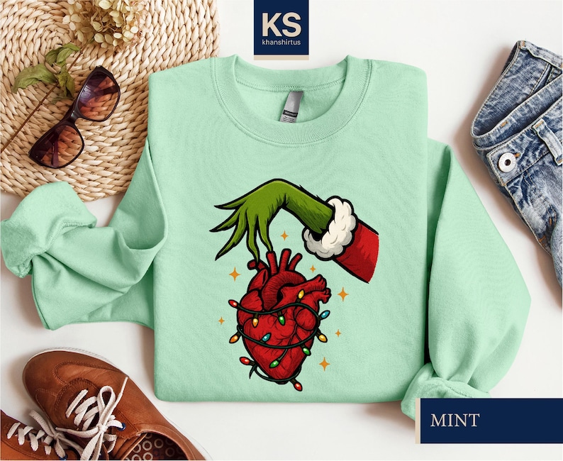A mint green sweatshirt with a graphic of a hand holding a heart with lights, a Santa Claus hand, and a Christmas tree. The sweatshirt is laid out on a white surface, along with a pair of sunglasses, a pair of jeans, and a pair of brown shoes.