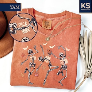 May include: Orange Comfort Colors t-shirt featuring a dancing skeleton design with a pumpkin and bats. The shirt has the text "FUCK TRUMP" in a circle. A gold necklace and a brown hat are also visible.