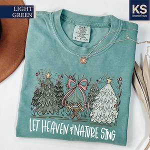 May include: Light green t-shirt with a Christmas-themed graphic. The design includes illustrated Christmas trees, a pink bow, and the text "LET HEAVEN & NATURE SING". A gold necklace is draped across the shirt.