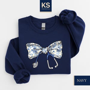 May include: Navy blue sweatshirt with a white and blue floral bow and a stethoscope design. The sweatshirt has a crew neck and long sleeves.