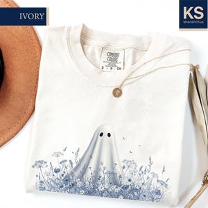 a white t-shirt with a ghost design printed on it, along with a hat, a necklace, and some dried leaves.