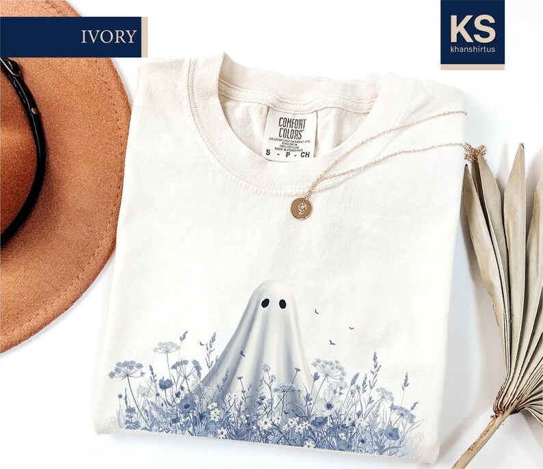 a white t-shirt with a ghost design printed on it, along with a hat, a necklace, and some dried leaves.