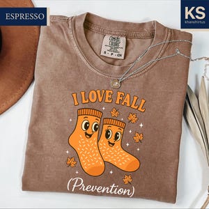 May include: A brown Comfort Colors t-shirt with the text "I LOVE FALL" and a graphic of two orange socks with smiling faces. The word "Prevention" is below the graphic. A silver necklace is draped across the shirt.