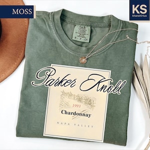 a green t-shirt with a wine label design, a brown hat, and some dried leaves, all placed on a white background.
