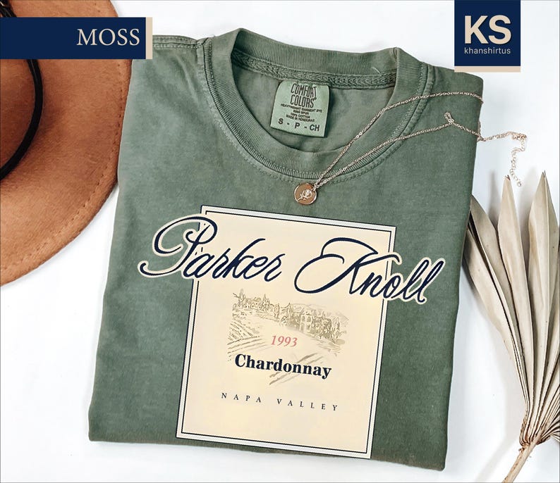 a green t-shirt with a wine label design, a brown hat, and some dried leaves, all placed on a white background.
