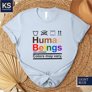 May include: Light blue t-shirt with a graphic of a washing instruction symbol, the word "Human" in brown, "Beings" in rainbow colors, and the text "100% Organic, Colors may vary."
