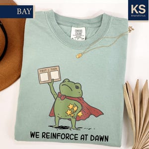 May include: A bay-colored t-shirt featuring a graphic of a green frog wearing a red cape, holding a sign that reads FIRST THEN and a cluster of yellow stars. The text WE REINFORCE AT DAWN is printed below the frog. The shirt has a crew neckline.