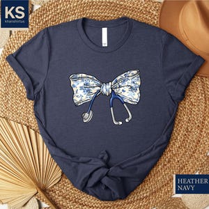 May include: A navy blue t-shirt with a floral print bow and a stethoscope. The text "HEATHER NAVY" is printed on the bottom right corner of the image.