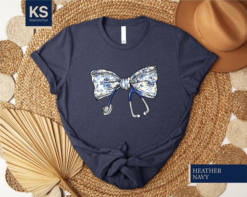 May include: A navy blue t-shirt with a floral print bow and a stethoscope. The text "HEATHER NAVY" is printed on the bottom right corner of the image.